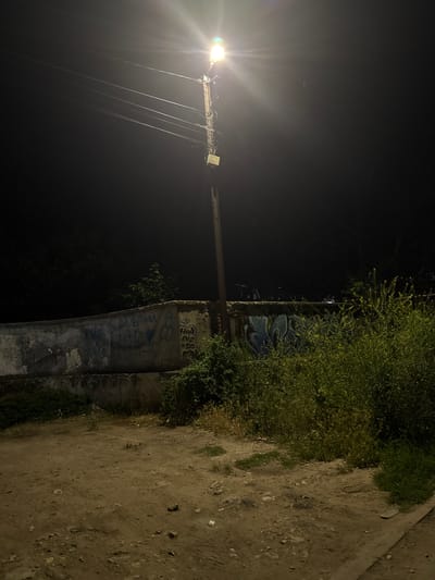 A street lamp illuminates a dimly lit outdoor scene at night, casting a bright glow and lens flare upwards. The lamp is attached to a wooden pole with visible wires. In the foreground, a dirt path with scattered debris leads towards a low, weathered concrete wall covered in graffiti. Beyond the wall, there are dense green bushes and trees. The overall environment appears to be an unkempt, possibly abandoned area within the city of Lom, Bulgaria.