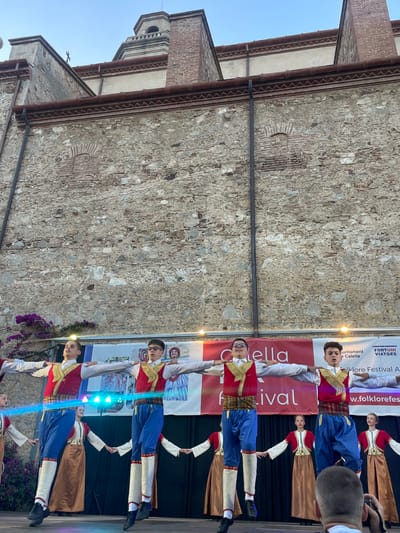 The image depicts a performance taking place outdoors, in front of a weathered stone wall and a building with red-tiled roofs. A group of young people, dressed in traditional folk costumes, are performing on a stage illuminated by stage lights. The costumes consist of red vests with gold trim over white shirts, blue pants, and white stockings with red accents. Some performers wear dark shoes.
The performers appear to be engaged in a dance or synchronized movement, with several individuals holding hands or extending their arms. The overall atmosphere suggests a festive event, possibly a folk festival. The lighting is artificial, indicating the performance is likely taking place in the evening or late afternoon.
In the background, there are banners with text. One banner partially visible contains the text "Calella Festival". Another banner has text that appears to be in Catalan, including "Ajuntament de Calella" and "folklorefestival". The presence of these banners and the mention of "Calella" in the context of a festival strongly suggest the location is Calella, Spain. Purple flowers are visible near the bottom left of the frame. The sky is clear and appears to be a pale blue, suggesting fair weather. The emotions conveyed are primarily focus and engagement from the performers.