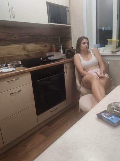 A young woman is sitting in a chair in a kitchen. She is wearing a white tank top and pink and white patterned shorts. Her legs are crossed, and she is looking off to the side with a neutral expression. The kitchen has light-colored cabinets and a wooden backsplash. There is a microwave above the backsplash, and a stovetop with a frying pan on it. In the foreground, on a white countertop, is a pack of Camel cigarettes and an ashtray with cigarette butts. The room appears to be dimly lit, suggesting it is evening. The location is identified as Mezhdurechensk, Russia.