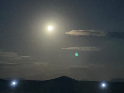 The image depicts a nighttime landscape scene. A bright, full moon is visible in the upper center of the frame, partially obscured by clouds. Below the moon, a distant mountain range is silhouetted against the twilight sky. Two bright lights, possibly streetlights or headlights, are situated on the horizon, illuminating the foreground. The overall atmosphere is serene and mysterious, with the moonlight casting a soft glow over the landscape. The specific location is the city of Kara-Oi, Kyrgyzstan.