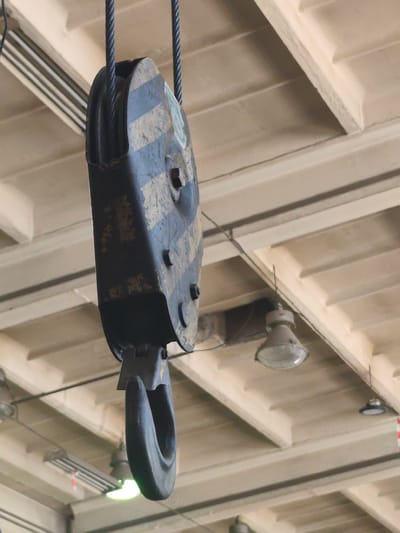 The image shows a large, industrial pulley system hanging from a ceiling. The pulley is made of dark metal with yellow and black stripes, and has a hook attached to its bottom. The pulley is suspended by two thick ropes. The ceiling appears to be part of a large, indoor industrial space, possibly a warehouse or factory, with visible light fixtures and structural beams. The lighting suggests it is daytime. There are no people visible in the image. The image focuses on the pulley system and its immediate surroundings.