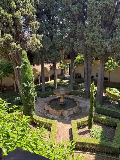 The image depicts a courtyard garden in Granada, Spain, likely within the Alhambra. A central circular fountain with a tiered water feature is the focal point, surrounded by geometric parterres of manicured hedges and pathways. Tall cypress trees and other mature trees with dense foliage are strategically placed, casting shadows on the garden. In the background, an arcaded walkway with white walls and columns suggests historical architecture. Several individuals are present: a group of people are seated on a bench in the shaded arcade, and another person is standing in the walkway, taking a photograph. The foreground is partially obscured by lush green foliage, suggesting the vantage point is from an elevated position. The sunlight indicates it is daytime.