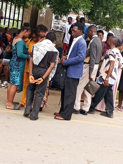 This media file captures a street scene in Guzape, Nigeria, with a group of people gathered, possibly waiting for something or observing an event. The subjects include several individuals, both men and women, dressed in a variety of casual and semi-formal attire. One woman in the foreground wears a shimmering blue dress, while another is in an orange outfit with smiley face motifs. Several men are seen in shirts and trousers, with one man in a blue blazer and another in a grey sweater.
The activity is unclear, but the individuals appear to be standing and conversing. Some are looking in the same direction, suggesting a shared focus. The background shows a wall with some posters, and behind that, a leafy tree canopy. There are also metal railings on the left, hinting at a fenced area or structure.
Notable details include the footwear of the people, ranging from sandals to dress shoes. The lighting suggests it might be daytime, with overcast conditions. The general atmosphere appears neutral, with no overt expressions of strong emotion visible.
There is some visible text on posters in the background, but it is largely illegible due to the resolution. One poster appears to have a phone number starting with "070..." and some images related to an event or service. The setting is an outdoor, paved area, likely a street or walkway, with a yellow line marking a boundary.