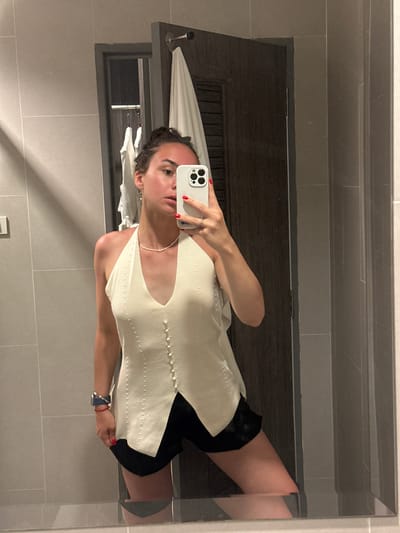 A person is taking a mirror selfie in a bathroom in Baan Klong Prao, Thailand. The individual is wearing a light-colored top with buttons down the front and dark shorts. A towel rack with towels is visible behind the person, along with a dark wooden door. The bathroom has light grey tiles on the walls.