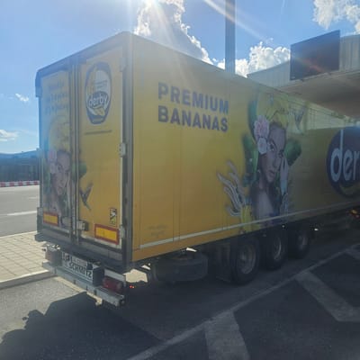 The image depicts a yellow truck with "PREMIUM BANANAS" printed on the side, along with a graphic of a woman wearing bananas and flowers in her hair, and a hummingbird. The truck also has a logo that reads "derby ECUADOR". The license plate reads "AE-329 SA" and "SCHMITZ" can be seen on the back. The truck is parked next to a building with a sign showing a red "X". The location appears to be Sid Municipality, Serbia.