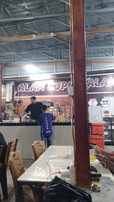 This is a medium shot photograph taken inside a casual eatery, likely a coffee shop or warung, in Keude Trienggadeng, Indonesia. The setting appears to be an indoor space with a corrugated metal roof and exposed beams, suggesting an open-air or semi-outdoor structure. A large wooden pillar dominates the right side of the frame.

In the background, a counter displays various items, including glasses and what appear to be coffee-making equipment. Two men are visible behind the counter: one in a dark t-shirt, appearing to be the barista or owner, and another younger person wearing a blue shirt with "REBORN CLUB" printed on it. The younger person is facing away from the camera, looking towards the counter.

The overall scene suggests a relaxed atmosphere, possibly during daytime given the lighting, though it's difficult to determine the exact time. The tables in the foreground, some empty and one with a white surface and what looks like lettering or reflections, are set up for customers. The presence of electrical outlets and charging cables on the table indicates that patrons may be working or socializing while using their devices. Red plastic containers are visible in the background, possibly for storage or as coolers. Text visible in the image includes "ALAM KUPI ALA", "AKABIKA", "NEW YORK, NY", "CEE", and logos from "HSG Fashion" and "HERISON". The image conveys a sense of everyday activity at a local establishment.