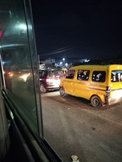 At night in Owode-Elede, Nigeria, the scene depicts road traffic. A yellow commercial van, commonly used as a passenger vehicle, is adjacent to a dark SUV. The photograph appears to be taken from inside another vehicle, as a window frame is visible. The road surface is asphalt, and distant building lights are seen in the background.