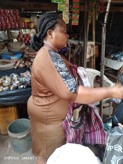 At an open-air market, a woman in a brown dress with shimmering sleeves stands beside a table laden with goods, including dried goods in plastic bags and canned items. A bucket sits on the ground nearby. Behind her, a white plastic chair is partially visible amidst the market's framework. The foreground is occupied by a white surface, and a black shopping bag with white lettering is held by an unseen hand. The overall scene is busy and indicative of a marketplace setting.