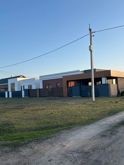 The scene depicts a clear, sunlit daytime view of a residential area in Dmitrievskaya, Russia. In the foreground, an unpaved dirt road runs horizontally, leading to a sprawling, mostly green and brown grassy field that extends to a row of modern-style single-story or low-profile houses. No individuals are present in the image.

Prominently featured in the mid-ground is a light gray concrete utility pole positioned slightly right of center. It extends from the bottom of the frame to near the top, equipped with an antenna or mast at its peak, a small white utility box, and a surveillance camera or sensor directed towards the foreground. A single, dark overhead utility line sags horizontally across the upper third of the image, originating from this pole and extending leftwards out of frame.

Three houses are visible, oriented horizontally across the mid-ground. The closest and most detailed house, on the right, exhibits a contemporary design with a flat roofline. Its facade combines reddish-brown brick, white horizontal banding, dark brown/bronze accent panels, and dark gray or stone cladding on its right side. A numerical marking, "M 399," is visible on the brown facade. This house is fronted by a dark gray horizontal slat fence, interspersed with stone-clad pillars. A second, similar modern house, predominantly white with dark-framed windows, is situated to its left, partially obscured. Further left, a third house, also white with a dark gabled roof, appears to be under construction or recently completed, with scaffolding-like elements on its leftmost side.

Several small, slender, leafless saplings are sparsely planted in the grassy area directly in front of the houses. The sky above is clear and bright blue, devoid of clouds. The overall lighting suggests either early morning or late afternoon, characterized by long shadows and a golden hue on some building surfaces.