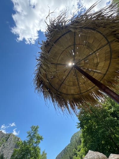 An upward-angle shot reveals a large, thatched umbrella casting a shadow under a bright blue sky with scattered white clouds. The umbrella's straw material is worn and frayed, with strands extending outwards. Sunlight pierces through gaps in the thatch, creating bright spots. A thick, dark brown pole supports the umbrella, angled upwards. In the background, lush green trees and a rugged, grey mountain range are visible. The scene suggests a natural, possibly recreational area.