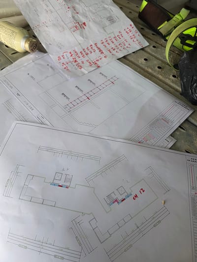 The image shows a cluttered workspace with several construction-related documents, a yellow and black safety harness, and a bottle of Maxim coffee. The documents are blueprints or technical drawings, with some handwritten notes and calculations in red ink on one of the pages. One drawing appears to be a floor plan with room designations like "CH 12". Another document has columns with labels such as "수직재" (vertical member) and "안전발판" (safety plank), suggesting it relates to scaffolding or construction safety.
The scene is likely an outdoor or semi-outdoor construction site, given the presence of the safety equipment and the general disarray of the papers. The documents are spread out on a metal surface with circular perforations, possibly part of the scaffolding itself or a temporary work platform. The lighting suggests it is daytime.
The handwritten notes on one of the documents include measurements, quantities, and Korean phrases like "대각재" (diagonal brace), "자동·고정 클램프" (automatic/fixed clamp), and "망, 발끝막이" (net, toe board), all pertaining to scaffolding. The coffee bottle indicates a break or a moment of work in progress. The text "311동-지붕층" (Building 311 - Roof Floor) and "PROJECT TITLE 대전" (Project Title Daejeon) provides specific location context within Daejeon, South Korea. The overall impression is one of active construction planning or site work.
