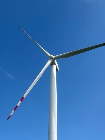 This media file is a low-angle shot of a wind turbine against a clear blue sky. The scene is dominated by the immense white blades of the turbine, which are angled upwards, with one blade extending horizontally across the frame. The tips of the blades are marked with red and white stripes. The sky is a vibrant, unblemished blue, suggesting a bright, clear day, likely during daylight hours. There is a hint of a thin white cloud on the horizon. The image focuses solely on the wind turbine and the sky, with no other objects or signs of human presence visible. The perspective from below emphasizes the scale and engineering of the wind turbine, while the clear sky suggests a peaceful and potentially windy environment suitable for energy generation. There is no visible text in the image. The location context of Abrykosivka, Ukraine, implies this is likely part of a wind farm in a rural or semi-rural setting.