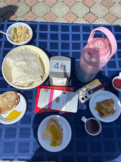 On a table covered with a dark blue checkered tablecloth, there is a picnic spread. The setting is outdoors, as indicated by the dappled sunlight and shadow patterns on the checkered pavement in the background, which appears to be a pathway in the city of Duvankul', Russia.

In the upper left corner, a white plastic bowl contains a portion of rice pilaf with visible pieces of meat and carrots, accompanied by two dill pickle spears and a plastic fork. To the right of the pilaf, a woven basket holds a folded cream-colored fabric, possibly a tablecloth or napkins, with an embossed floral pattern. Next to the basket is a gray dispenser with the number "40" and a small circular logo featuring a cartoonish figure.

Dominating the upper right side of the image is a large, transparent pink water bottle with a handle, marked with time-based motivational hydration reminders. A small white cup filled with a dark red liquid, likely tea or juice, is positioned to the right of the water bottle.

Below the water bottle, a smartphone is laid flat, with keys and a brown leather keychain attached to it. To the right of the phone, a white plate holds a folded pancake topped with a yellow sauce or jam. Another white plate in the lower left displays two pancakes, one on top of the other, also with a yellow topping. A red and silver package of "BUCHERON CHOCOLATE" is visible in the center of the arrangement, with Russian text visible on the bottom part of the packaging. A small white cup, similar to the one holding the red liquid, is placed below this package and to the left of the pancake plate.