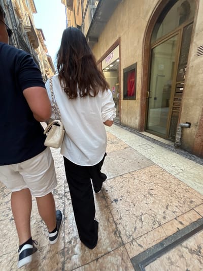 The image shows a street scene in Verona, Italy, featuring two individuals walking on a paved street. The person in the foreground is wearing a dark blue shirt and light-colored shorts and black/white sneakers. The person in the background has long brown hair, wearing a white shirt and black pants, and a small white bag with a chain strap. The background shows buildings and shopfronts typical of Verona, including a "KIKO" store. The setting is urban, with architectural elements visible in the background buildings.