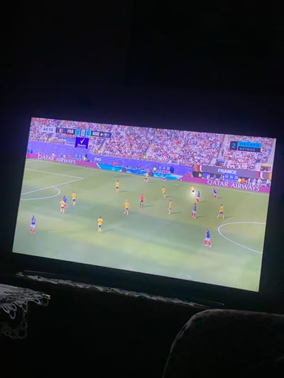 The image shows a television screen displaying a soccer match. The score is 0-0 between France (FRA) and Sweden (SWE) at 44:10. Players in blue and yellow jerseys are on the green field, with a large crowd visible in the stands. Advertising boards for Qatar Airways and FIFA are present around the field. The screen is angled and slightly blurry, suggesting it is being viewed from an off-center position. The environment around the television is dark, with the lower part of the image showing a glimpse of dark furniture and what appears to be a lace doily. This scene is in the city of Zastavna, Ukraine.