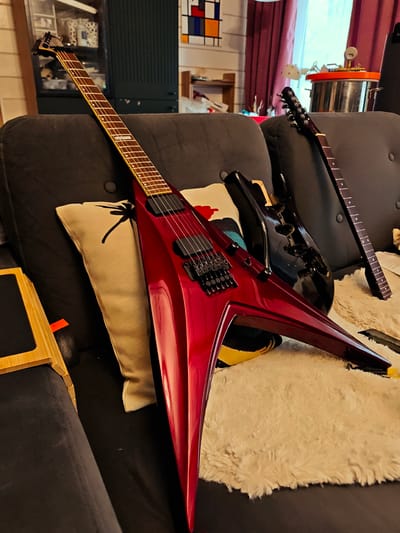 The image depicts a domestic interior, likely a living room, though the specific location in Свистуха (кузяевский С/о), Russia, is not verifiable from the visual data alone. Dominating the foreground is a red, V-shaped electric guitar leaning against a cushion on a dark grey sofa. Another black electric guitar body, seemingly disassembled, is partially visible behind the red guitar. A guitar neck is lying on a fluffy white rug on the sofa seat. In the background, there is a cabinet, a Mondrian-style artwork, and other household items. The room's lighting appears natural. No people are visible in the frame.