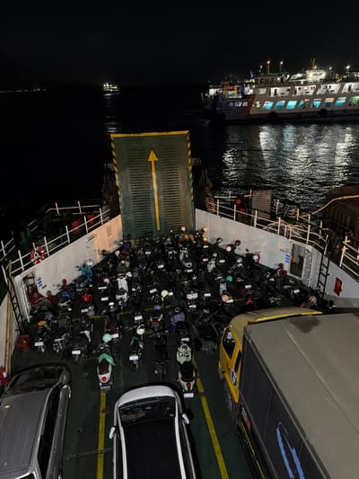 This high-angle night shot captures a ferry docked at the city of Ketapang, Indonesia, loaded with vehicles and motorcycles. The ferry's deck is densely packed with numerous motorcycles, most of which are dark-colored. Two cars, one silver and one white, are also visible on the deck. A yellow truck with a gray cargo container is parked near the right edge of the frame. Several people are present on the ferry. Two individuals are sitting near a life preserver on the left side of the ferry. The ferry is illuminated by artificial lights, reflecting on the dark water surrounding it. In the background, another ferry is visible, also illuminated and navigating through the water. The scene depicts a typical night operation for vehicle transport by ferry in the region.