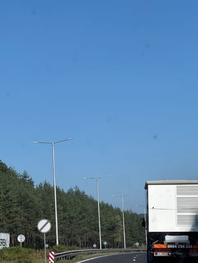 This media file captures a roadside scene under a clear blue sky. The primary focus is a section of a highway with a dense line of evergreen trees bordering it. Several tall, modern streetlights with horizontal arms extend over the road. A large white truck with a corrugated side is parked on the right side of the road, partially visible. The truck has a license plate that reads "CO 3485 CT" and a phone number "0884 734 335" printed on its side. There are also several road signs visible, including a circular sign with diagonal black bars on a white background, indicating the end of a restriction, and a red and white striped sign that likely denotes a hazard or warning. The lighting suggests it is daytime, possibly late morning or afternoon, with no indication of clouds or adverse weather. The image appears to be taken from a moving vehicle or stationary on the side of the road, looking towards the highway and the adjacent forest. The overall impression is one of a typical rural highway scene in Bulgaria, specifically in the Dolna Vasilitsa area, characterized by infrastructure for road lighting and traffic management, flanked by natural greenery.