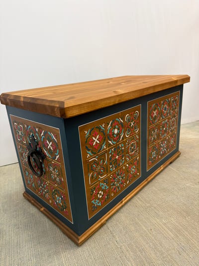 The image shows a decorative wooden chest with a natural wood lid. The sides of the chest are painted a dark teal, and feature panels decorated with a mosaic of colorful, intricate tile-like patterns in shades of red, blue, white, and brown. A dark metal ring handle is attached to the front left side of the chest. The chest is placed on a textured, light-colored surface, possibly a rug or carpet. The background is a plain white wall, suggesting an indoor setting. There is no indication of the city of Maloyaroslavets, Russia, or any other specific location.