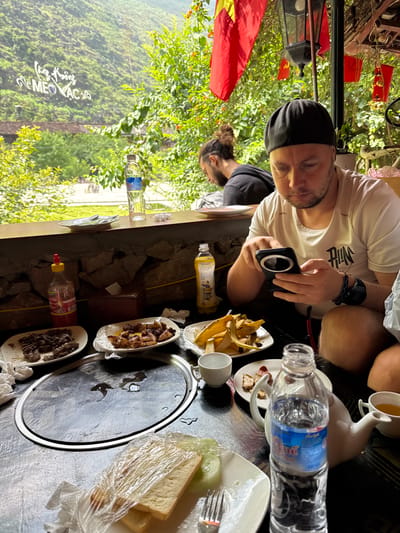 The image shows a man in a white shirt and black cap seated at a table outdoors, holding a black electronic device. A second man with a man bun is visible in the background, slightly out of focus. The table is laden with food, including plates of meat, fries, bread, and various bottles and containers. A circular cooking surface sits in the center of the table. The scene appears to be in a mountainous area, identifiable as Mèo Vạc Commune, Vietnam, based on the distinctive landscape visible through the open-air structure. A Vietnamese flag is also visible in the background.
