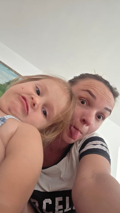 The media file shows a close-up, upside-down selfie of two people, likely a mother and child, in an indoor setting. The person in the foreground is an adult woman with dark hair, brown eyes, and prominent forehead wrinkles. She is wearing a white t-shirt with black stripes on the sleeves and what appears to be text on the front that is partially visible, reading "CEL". She is sticking her tongue out, with a playful expression.
Behind her, and slightly above, is a young child with blonde hair and brown eyes. The child is looking towards the camera with a curious expression and has their mouth slightly open. They are wearing a blue and white patterned garment. The background is a plain, light-colored wall, suggesting an indoor environment. A portion of what looks like a framed artwork with a green and blue landscape can be seen in the upper right corner, above the child's head. The lighting appears to be bright, indicating it could be daytime. The overall mood of the image is lighthearted and candid, capturing a moment of playful interaction between the two subjects. The location context provided is Guía de Isora, Spain, which does not offer further visual cues in the image itself.