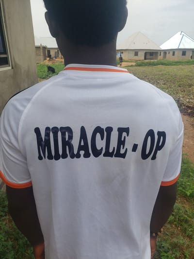 A close-up, rear-facing shot shows the back of a person wearing a white t-shirt with "MIRACLE - OP" printed in dark blue lettering. The t-shirt has orange trim around the collar and sleeves. The background suggests an outdoor environment, possibly a rural or semi-urban area, with grassy terrain and buildings under construction. In the distance, two unfinished structures with distinct rooflines are visible. One building has a grey, pitched roof, while the other features a white, peaked roof. A dark-colored animal, possibly a goat, is present in the middle ground, near the buildings. A person is also visible in the far distance, standing near the construction site. The overall scene appears to be in Kachia, Nigeria, given the architectural style and vegetation.