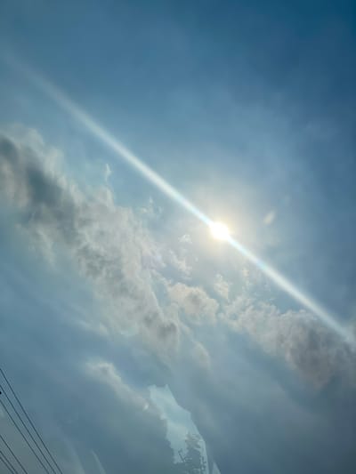 The image is a low-angle shot of a bright sky with scattered clouds. The sun is a prominent, luminous disc, casting a strong diagonal ray of light across the frame. The sky transitions from a pale blue at the top to a slightly darker blue towards the bottom. Some thin, dark power lines are visible in the lower left corner, suggesting an outdoor setting. The image does not provide any specific details or context that would allow for the identification of the city of Mandar Sari, Indonesia, or any inhabitants.