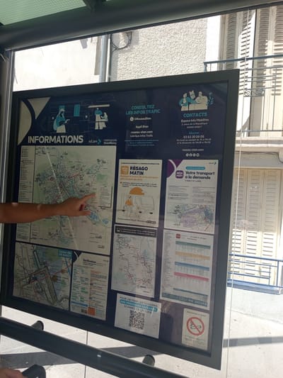 The image shows a close-up view of a public transportation information board located in Nancy, France, likely at a bus stop or transit station. The board is large and displays various maps, schedules, and contact information for the local transit system, Stan, and the Grand Nancy metropolitan area.
In the foreground, a person's left hand and forearm are visible, with the index finger pointing towards a section of a bus route map displayed on the board. The hand is the only visible part of a person.
The information board itself is the central subject. It is divided into several sections. On the left, a large map of Nancy and its surrounding areas shows bus lines, tram lines, and other points of interest. To the right of the map, there is information about consulting traffic updates, contacts for information mobility services, and a section titled "Vos transports à la demande" (Your on-demand transport). Further to the right, there are more detailed route maps and schedules for specific bus lines, such as "C2 LAXOU Pl. de Haye - Vandoeuvre - Nancy." At the bottom right of the board, there is a "NO SMOKING" sign with a cigarette crossed out.
The environment appears to be outdoors, indicated by the natural lighting and the buildings visible in the background. The buildings have a textured facade and shutters, typical of French architecture. A metal railing is visible in the lower part of the frame, suggesting it's part of the bus stop or station infrastructure. The overall scene is bright, suggesting it was taken during daylight hours.