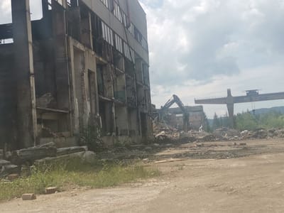 The image depicts an industrial demolition site in Zhivkovo, Bulgaria, under an overcast sky during daylight hours.
**Subjects:** The primary subject is a large, multi-story industrial building, likely a former factory or warehouse, that is in an advanced state of partial demolition and decay. Its concrete and brick facade shows significant damage, with numerous broken windows, exposed structural elements, and missing wall sections. In the mid-ground, a large, dark-colored excavator is visible, with its arm raised, surrounded by extensive piles of rubble consisting of broken concrete, twisted metal, and other debris. Further in the background, a massive, weathered gantry crane structure stands prominently against the cloudy sky. Distant green mountains and scattered trees form the horizon line.
**Activity or event taking place:** The scene clearly illustrates an ongoing or recently completed demolition event. The excavator's presence and the vast amount of debris confirm that the industrial building is being dismantled.
**Notable details:** The weather is cloudy, with a bright, diffused light suggesting it is daytime. The ground in the foreground is a dirt path with patches of green grass and scattered bricks. The decaying structure of the building, with its exposed inner workings and broken glass, emphasizes the destructive nature of the activity. The gantry crane provides a strong visual cue to the site's previous industrial function. The general atmosphere suggests a transition from an active industrial zone to a cleared or redeveloped area.
**Visible text:** The word "HYUNDAI" is visible on the arm of the excavator.