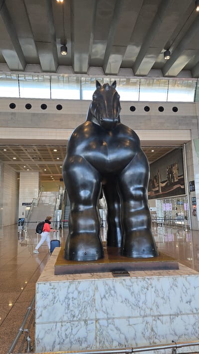 A large, dark bronze sculpture of a horse, characterized by exaggerated muscular forms, stands prominently in the foreground of a modern indoor public space. The sculpture is mounted on a tiered base, the lower portion of which is clad in white marble with grey veining. The background reveals an airport terminal environment. To the left, a woman wearing a coral jacket, white pants, and sneakers is walking away from the viewer, pulling a dark blue rolling suitcase and carrying a black backpack. She is positioned near a set of escalators and stairs. In the distance, a large mural or artwork is visible on the wall, depicting several figures. The space features high ceilings with exposed structural elements and lighting fixtures, and a reflective floor. The overall impression is that of a contemporary art installation within a busy transportation hub. This sculpture is located in Terminal 1 of Barcelona-El Prat Airport in El Prat de Llobregat, Spain.