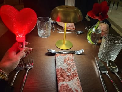 A dimly lit table setting features two place settings, each with a knife, fork, and spoon arranged around a patterned napkin. A gold-colored lamp illuminates the center, flanked by drinking glasses. One place setting includes a small vase holding a red tulip. A hand holds a glowing red heart-shaped object, adding a playful and romantic element to the scene.