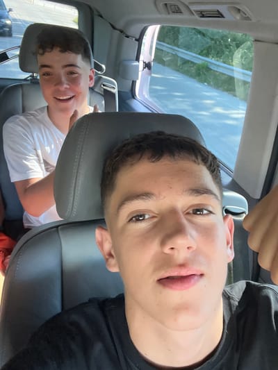 The image shows two young boys inside a vehicle. The boy in the foreground is positioned closer to the camera, with his head angled towards the viewer and his face filling most of the frame. He has short, dark hair and is looking directly at the camera with a slight smile. He appears to be wearing a black t-shirt.
Behind him, another boy is visible, seated in the back of the car. He is wearing a white t-shirt and smiling broadly, looking off to the side. The interior of the car is visible, including the grey leather seats and headrests. Through the car windows, greenery and sunlight suggest an outdoor setting, possibly a daytime scene. The angle of the sun and the shadows cast indicate it is likely late morning or early afternoon. The overall mood seems to be casual and lighthearted, possibly during a car ride. There is no visible text in the image.