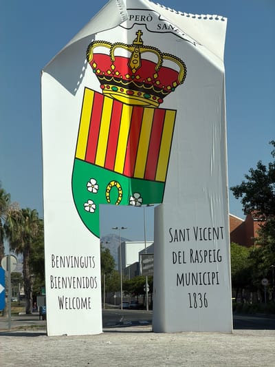 This is a full-shot, eye-level, outdoor photograph of a large, white, gateway-like structure with a banner that appears to be made of paper. The banner is curved and has a torn, perforated edge along the top. It depicts a coat of arms with a crown, a shield with red and yellow stripes over green, a horseshoe, and three white flowers. Text on the banner reads "PERO SANET".

The gateway structure is a monumental archway. The left pillar of the archway displays text in three languages: "BENVINGUTS", "BIENVENIDOS", and "WELCOME". The right pillar of the archway displays text in Spanish: "SANT VICENT DEL RASPEIG" and "MUNICPI 1836". The scene is set in a city environment. In the background, there are palm trees, buildings, streetlights, traffic signs, and mountains. The sky is clear and blue. The ground in front of the gateway is paved with light-colored aggregate material. There are no people visible in the immediate foreground or interacting with the gateway. The overall impression is of an entrance or a landmark in the city of San Vicente Del Raspeig, Spain.