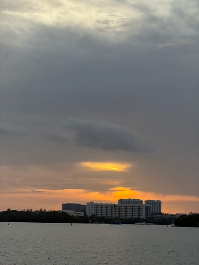 A view from the water looking towards a city skyline at sunset. The sky is filled with dark, ominous clouds with a bright, golden break in the clouds where the sun is setting. A line of trees and several tall buildings form the cityscape. Boats are visible on the water. The scene is located in Miami Beach, Florida, United States of America.