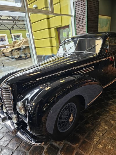 A black automobile, possibly a vintage model, is on display, likely indoors, as evidenced by the cobblestone floor and the reflection in its shiny surface. The image is slightly tilted, showing the front of the car with a large, curved fender, a spoked wheel, and chrome detailing on the grille and bumper. A display case with museum exhibits is visible through a window behind the car, along with other objects and photographs. The car's location is in Архангельское, Russia.