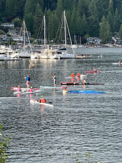 The image shows a waterfront scene in North Vancouver, Canada, with many sailboats docked in a marina in the background. The foreground and midground feature numerous individuals engaged in water activities on the lake, including kayaking and paddleboarding. Several people are in kayaks, some of whom are wearing life vests. Others are on paddleboards, with one individual standing and another sitting. The water is calm with visible ripples, reflecting the overcast sky. The shoreline is lined with dense evergreen trees and houses built on a hillside.