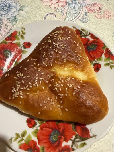 A golden-brown, triangular pastry, sprinkled with sesame seeds, rests on a white plate adorned with red poppies and green leaves. The plate is positioned on a tablecloth featuring a floral pattern with blue, green, and pink elements. The scene appears to be indoors, likely in a home setting. No specific location in Turinsk, Russia, can be determined from the image.