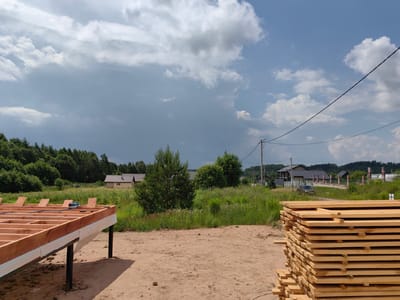 The image depicts an outdoor scene in the village of Garanski, Belarus, during daytime under a partly cloudy sky. In the foreground, a stack of lumber is prominently displayed on the right side. To the left, the wooden frame of a building foundation is visible, elevated on dark metal posts. The ground consists of sandy soil. In the middle ground, a lush green field with scattered wildflowers stretches towards a line of trees. Several houses with brown roofs are nestled amongst the trees. In the background, a dense forest borders the horizon, and utility poles with power lines are present. The overall atmosphere suggests a rural construction site.
