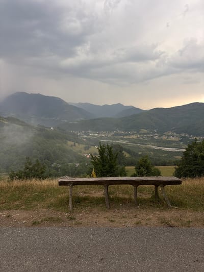 This image depicts a scenic view from Mekića Kula, Montenegro. The foreground features a rustic wooden bench placed on a grassy embankment next to an asphalt path. The bench appears weathered, with a simple design. Beyond the bench, the landscape unfolds into a valley with a winding river, dotted with a village that has houses with red and white roofs. The valley is surrounded by lush green, tree-covered mountains, which fade into hazy, layered peaks in the distance. The sky is overcast with dramatic, thick clouds, suggesting either early morning or late afternoon light, with a faint yellow glow towards the horizon. A light haze or mist is visible in the lower part of the valley, adding to the atmospheric quality of the scene. The overall mood is tranquil and contemplative, inviting the viewer to pause and appreciate the natural beauty of the mountainous region. There is no visible text in the image.