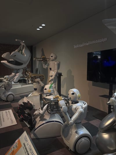 The image depicts an indoor exhibition featuring several white, humanoid robots with musical instruments, likely in the city of Nagoya, Japan. In the foreground, a robot with a large tuba-like instrument is positioned prominently. To its left, another robot holds a smaller trumpet, and further back, a third robot plays a trumpet. A larger, more complex robot structure is visible on the left side of the image, partially obscuring the background. In the background, a large screen displays reflections of people. The robots are arranged on a checkered floor. A sign with Japanese text is visible in the lower left quadrant, and another sign with the text "TODAY FOR TOMORROW" is visible in the lower right. The overall atmosphere suggests a technology or robotics exhibition.