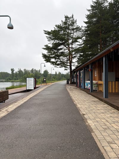 The image shows a paved walkway next to a body of water, likely the Aksakovka River, in Belebey, Russia. The walkway is lined with a wooden boardwalk on the left and a row of small shops or kiosks on the right. Pine trees are visible in the background. The sky is overcast, suggesting it may be a cloudy or rainy day. There are no people visible in the scene. The shops appear to be closed, and some have vending machines and refrigerators outside. A sign with Russian text is posted on the left side of the walkway. The scene depicts a public waterfront area with amenities for visitors.