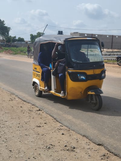 A yellow and black motorized tricycle, also known as an "okada," is captured in motion on an asphalt road. The tricycle, which appears to be a passenger vehicle, has a dark grey canopy. Inside the tricycle, two individuals are visible. The driver, wearing a dark blue garment, is seated at the front, operating the vehicle. A passenger, also in a dark blue garment, is seated behind the driver. A black bag is placed on the passenger's lap. The tricycle's side is marked with "DELUXE" and a license plate reading "BSR 2483". The road is bordered by a dirt shoulder with scattered stones. In the background, several buildings are under construction, constructed with concrete blocks and lacking roofs, suggesting an ongoing development or expansion in Kwamba, Nigeria. The sky is partly cloudy, indicating a day with mixed weather conditions.