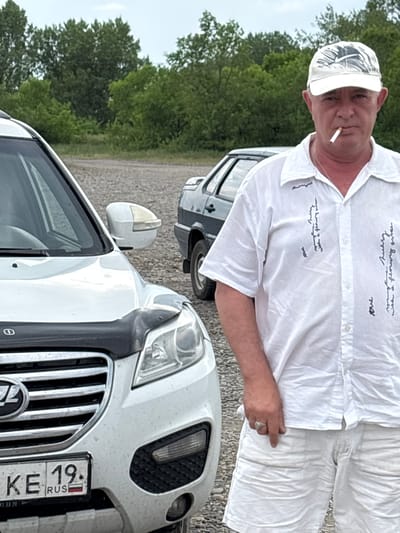 A man in a white shirt and shorts, wearing a white cap and smoking a cigarette, stands beside a white SUV. The man is looking at the camera. The SUV has a license plate that reads "KE 19. RUS". In the background, there are trees and another car, which appears to be a dark-colored sedan. The ground is covered with gravel. The overall setting suggests an outdoor, possibly rural or suburban location, in the city of Chernogorsk, Russia.