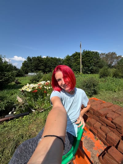 A young person with bright red hair takes a selfie in a sunlit garden. They are wearing a light blue t-shirt and dark grey leggings. Their left arm is extended, holding the camera. In the background, there are lush green trees, a greenhouse, and a patch of colorful lilies. To the right of the person, a pile of red bricks is visible. The sky above is a clear, bright blue. The location is the village of Zabolotovo, Russia.