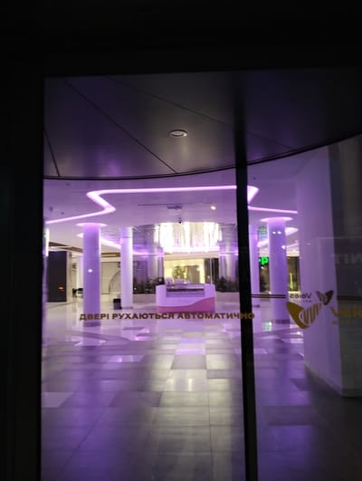 A view through a set of automatic doors reveals the interior of a mall. The sign on the doors reads "ДВЕРІ РУХАЮТЬСЯ АВТОМАТИЧНО", indicating that the doors open automatically. The text is in Ukrainian. The interior has a patterned tile floor, white columns, and a modern ceiling design with purple lighting. There's a kiosk in the center of the view, and other stores can be seen in the background. The sign on the window implies the location is in a shopping mall in Ivano-Frankivsk, Ukraine.