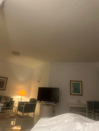 A dimly lit room features a wall with an indiscernible painting, a television mounted above a lit lamp, and a wall vent. An open-shelved cabinet is against the opposing wall, adorned with a framed print above it. A bed with white linens occupies the foreground, partially obscuring the view. Chairs are next to the cabinet.