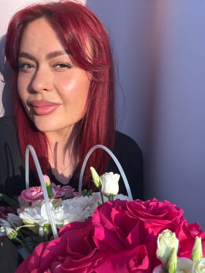 A close-up shot shows a young woman with bright red hair and freckles, smiling gently as she holds a bouquet of flowers. She is wearing a black top and a delicate silver necklace. The bouquet contains a variety of flowers, including large pink roses, white chrysanthemums, and pink carnations. The background is a plain, light purple wall, with a shadow cast from the woman's left side. The lighting suggests it is daytime, with sunlight hitting the woman's face from the right. The image is framed from the chest up, focusing on the woman's face and the colorful flowers. The location is unspecified in the image itself, but the prompt asks for it to be in the city of Узюково, Russia.