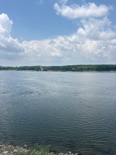 The image depicts a wide river with a dense, green forest lining the opposite bank. The sky above is blue with scattered white cumulus clouds. On the far bank, a large white construction crane is visible, suggesting ongoing industrial or development activity. The foreground shows a rocky shoreline with some sparse vegetation. The water surface is rippled, indicating a gentle breeze. The scene is serene and appears to be a natural landscape with a touch of human infrastructure. This location is likely along the Danube River in Silistra, Bulgaria.