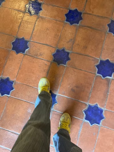 The image presents a first-person, downward perspective of a person's feet and lower legs standing on a distinctive tile floor. The individual is wearing bright yellow sneakers and dark-colored trousers, possibly corduroy or denim, which partially obscure the lower part of their legs.
The floor is a prominent feature, constructed from warm, reddish-brown terracotta-colored square tiles. These are laid out in a grid pattern. At various intersections or as decorative inserts within this grid are striking dark blue, eight-pointed star-shaped tiles. This combination creates an intricate and traditional mosaic pattern. The grout lines between the tiles are visible, defining the geometric layout. The tiles appear somewhat worn, indicating age and use, but are generally clean.
Subtle shadows are cast by the person's feet and legs, suggesting natural light from an overhead or side source, implying it is daytime. The architectural style of the tiled floor, with its characteristic terracotta and blue star motifs, strongly evokes traditional Spanish or Moorish design. This aligns perfectly with the given location context of Mérida, Spain, where such historical and culturally rich flooring is commonly found in old buildings, courtyards, or public areas. The image conveys a quiet moment of presence within a historically resonant setting. There is no visible text in the image.
