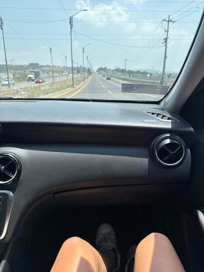 The image shows an interior view from the passenger seat of a vehicle, possibly a car, traveling along a highway near San Francisco Totimehuacán, Mexico. Outside the window are power lines and light posts lining the road, and there is evidence of other vehicles. Inside, a textured grey dashboard is prominent, with air vents visible. A portion of a person's legs, clad in light-colored clothing, and feet in grey shoes are visible on the right side of the image.